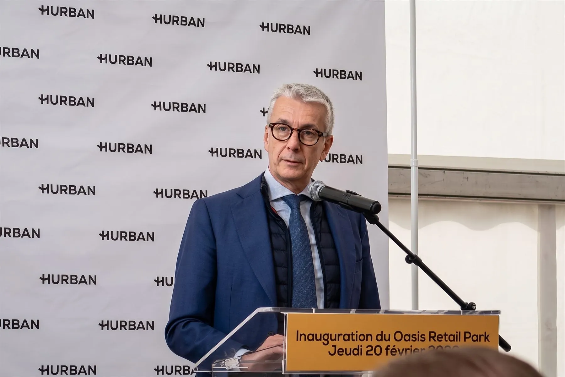 Prise de parole au pupitre pour l'inauguration de l'Oasis Retail Park, devant le mur de logos Hurban